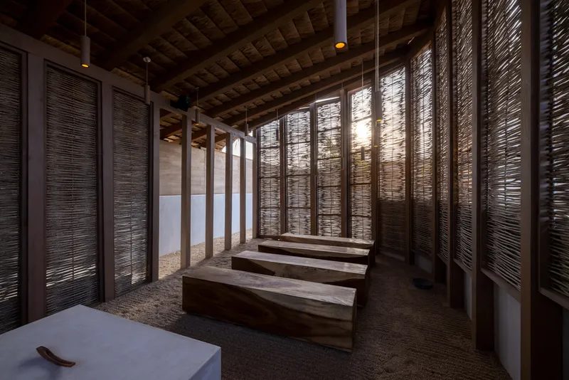 Timber benches arranged in rows beneath reed ceiling and backlit woven screen panels at dusk