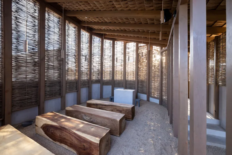 Interior with timber benches on gravel floor beneath exposed rafters and woven screen walls