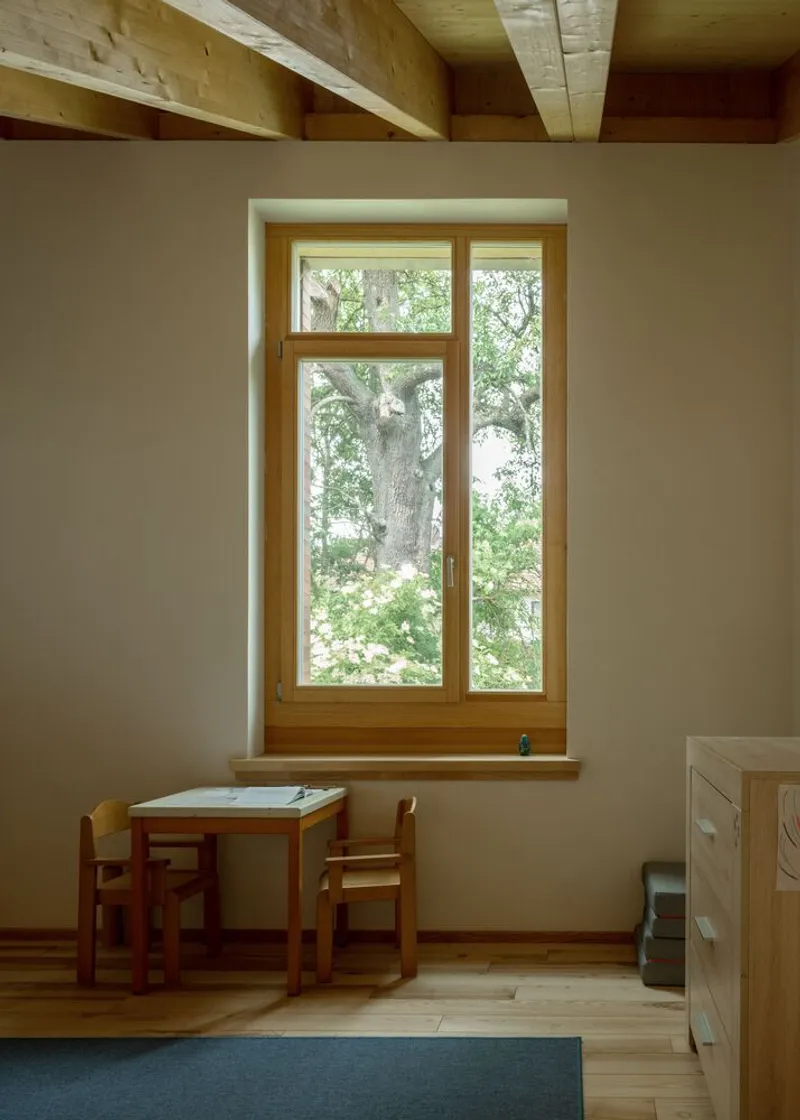 Tall timber-framed window with deep reveal overlooking trees, paired with a children's table