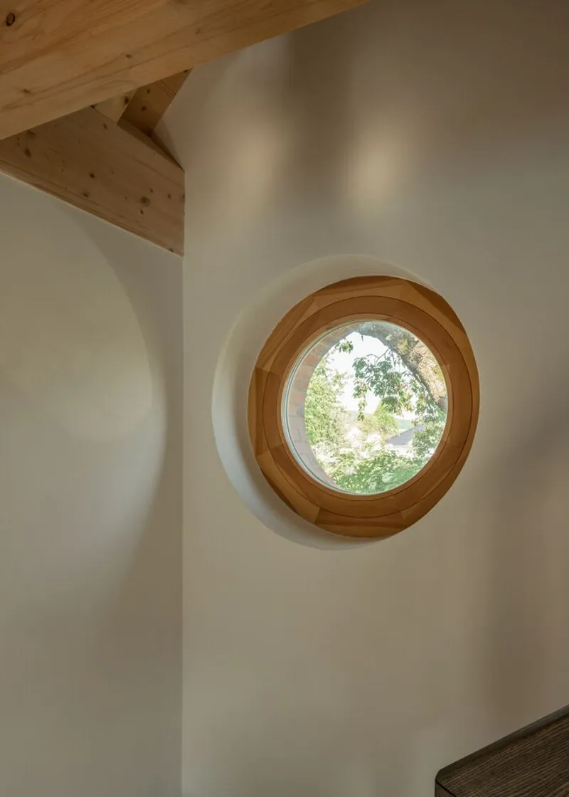 Close-up of circular timber-framed window set in a thick white wall below exposed rafters