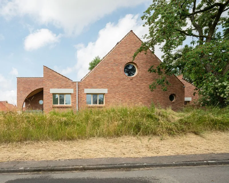 Red brick facade with gabled volumes and circular windows set behind a sloped grass berm