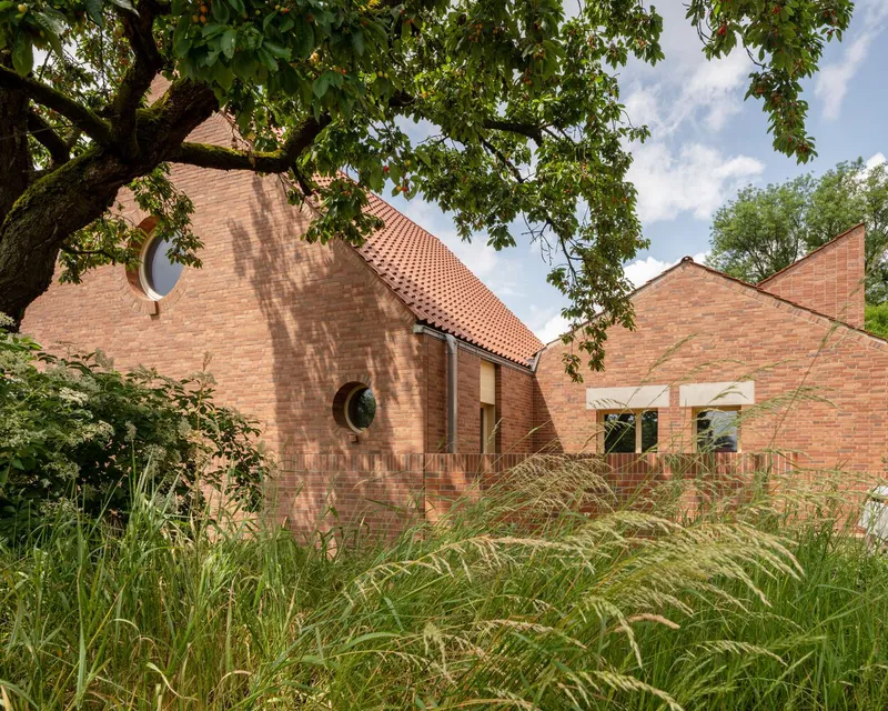 Brick gabled volumes with circular and rectangular openings framed by overhanging tree branches and tall grasses