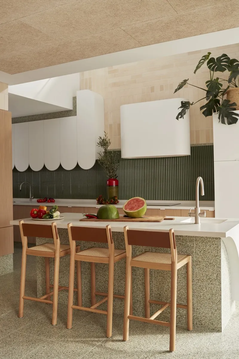 Kitchen island with terrazzo base and timber stools beneath a scalloped white range hood and cork ceiling