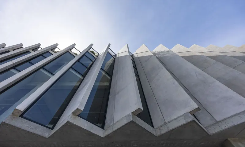 Upward view of zigzagging concrete facade with triangular skylights set against a clear blue sky