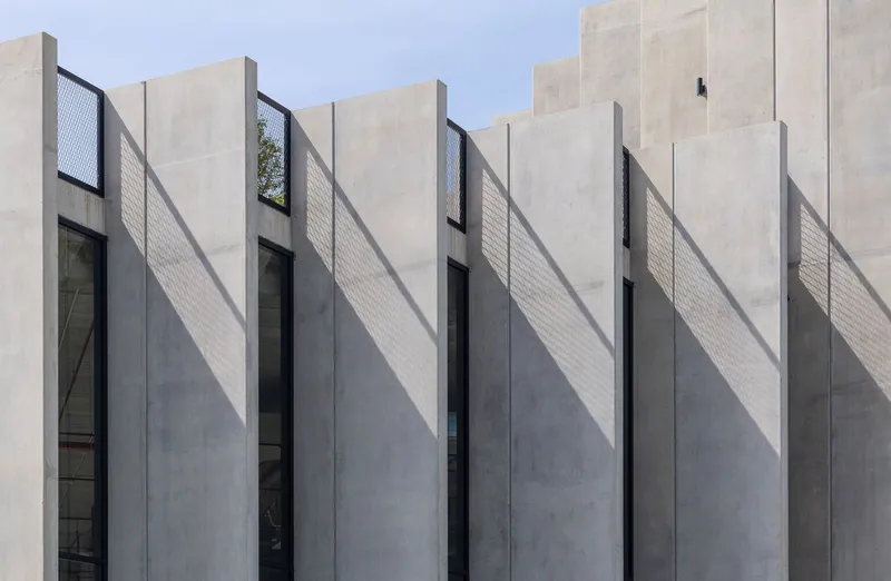 Concrete facade with vertical fins and mesh-screened openings casting diagonal shadows in bright daylight