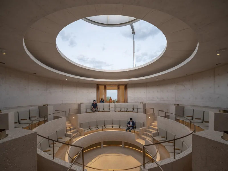 Interior circular sanctuary with tiered seating and central oculus opening to cloudy sky