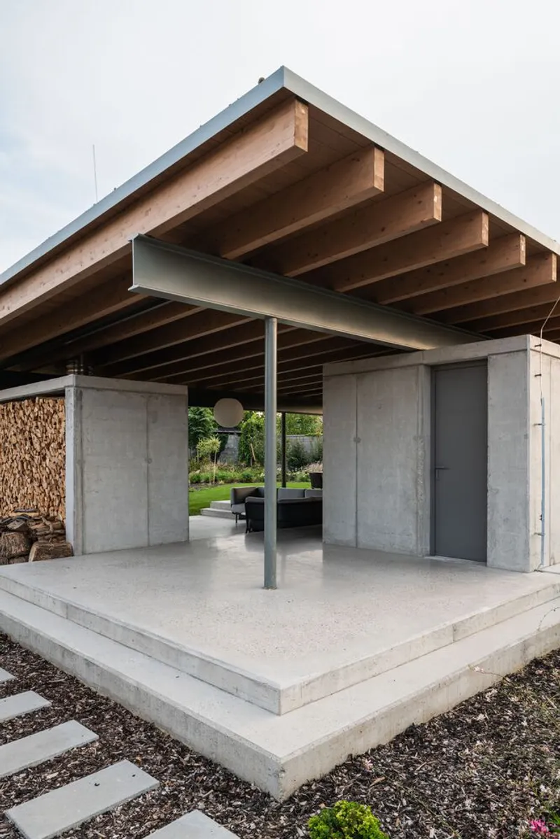 Covered outdoor terrace with exposed timber beams, concrete walls, steel column and stacked firewood under overcast sky