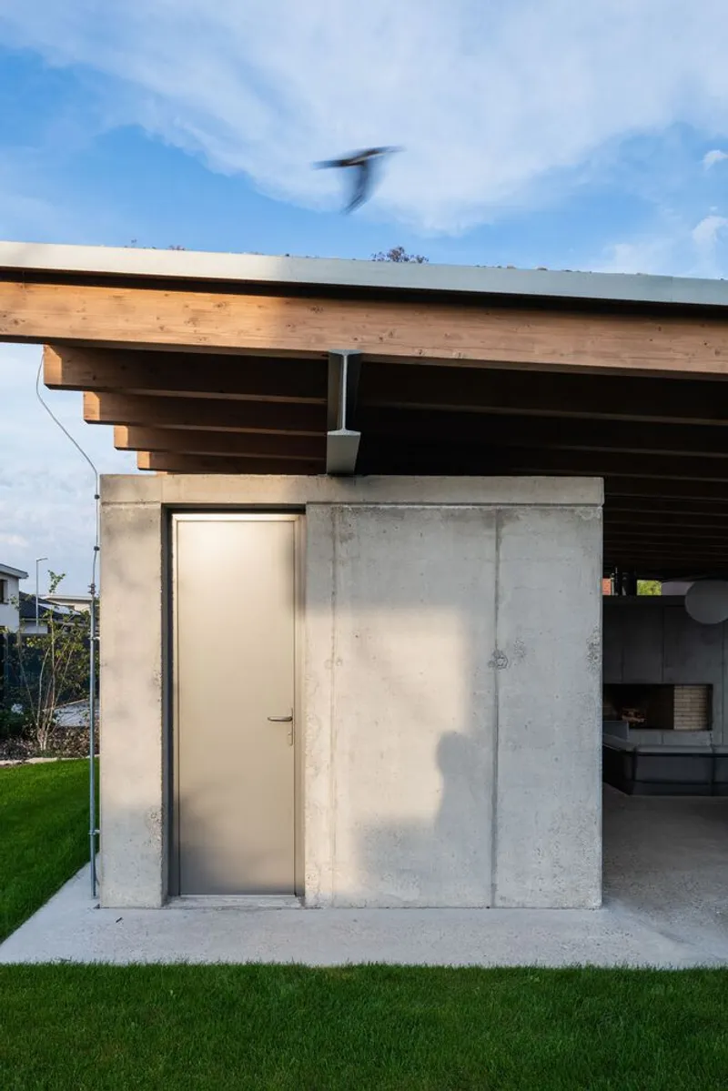 Concrete service volume with pale door beneath cantilevered timber beams and metal gutter against blue sky