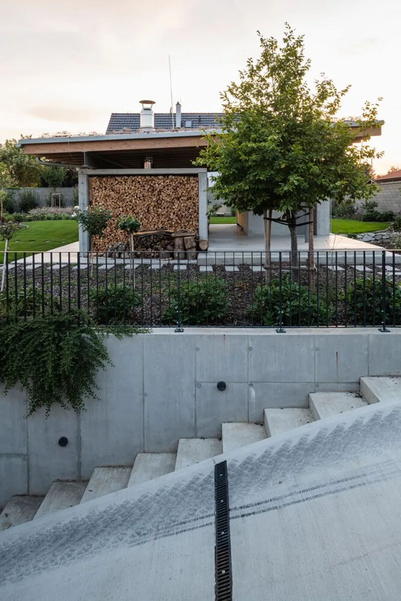 Concrete stairs descending to garden level with low walls, planted beds and pavilion with tree at dusk