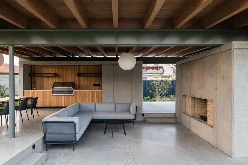Open pavilion interior with concrete fireplace, outdoor kitchen, timber cabinetry and sectional sofa under exposed joists