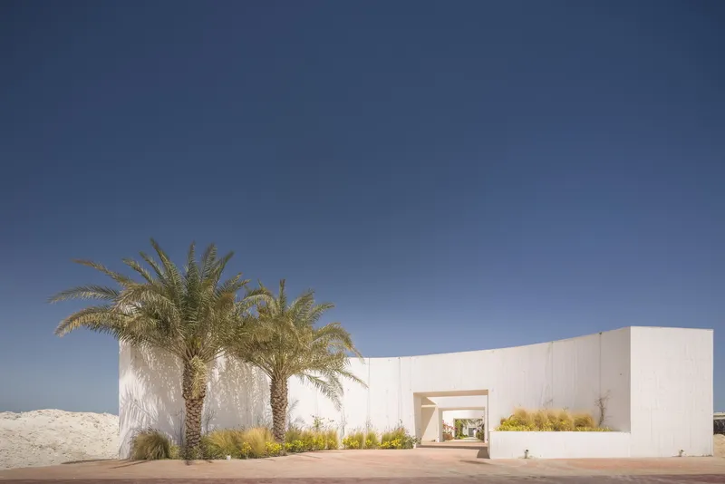 White rendered wall stretching across a sandy site with palm trees and grasses under a clear sky