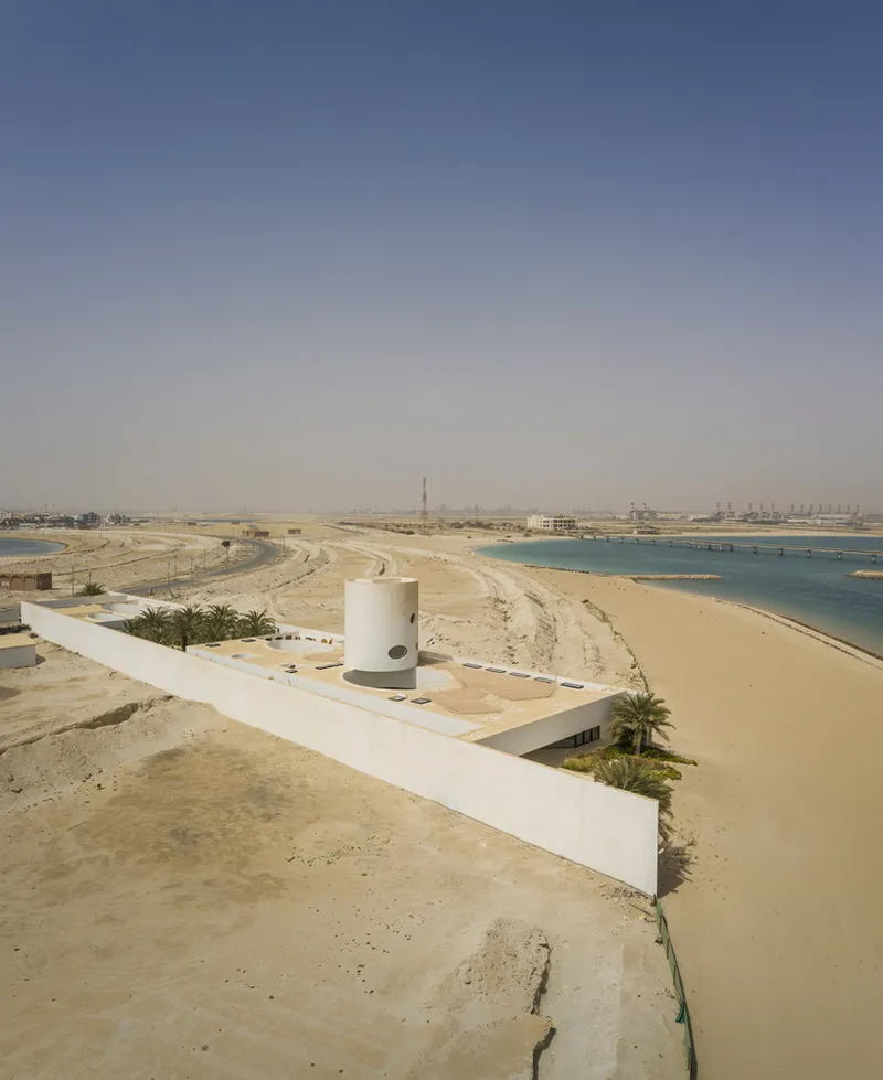 Elevated view showing the white linear structure and cylindrical tower set in the coastal sand landscape