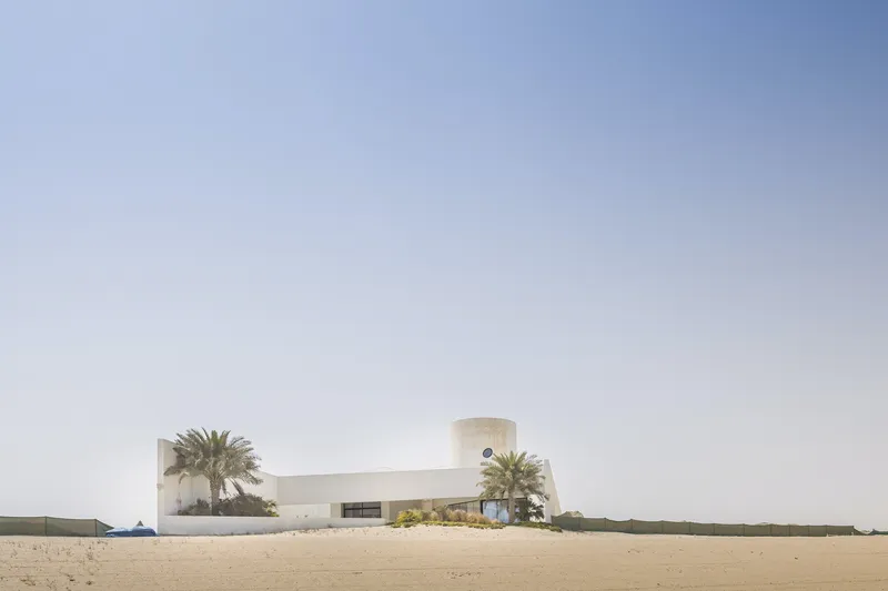 Distant view of the low concrete complex with cylindrical tower and palm trees in the desert sand
