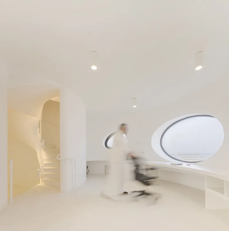 Interior hallway with arched openings, oval window, and blurred figure walking across terrazzo floor