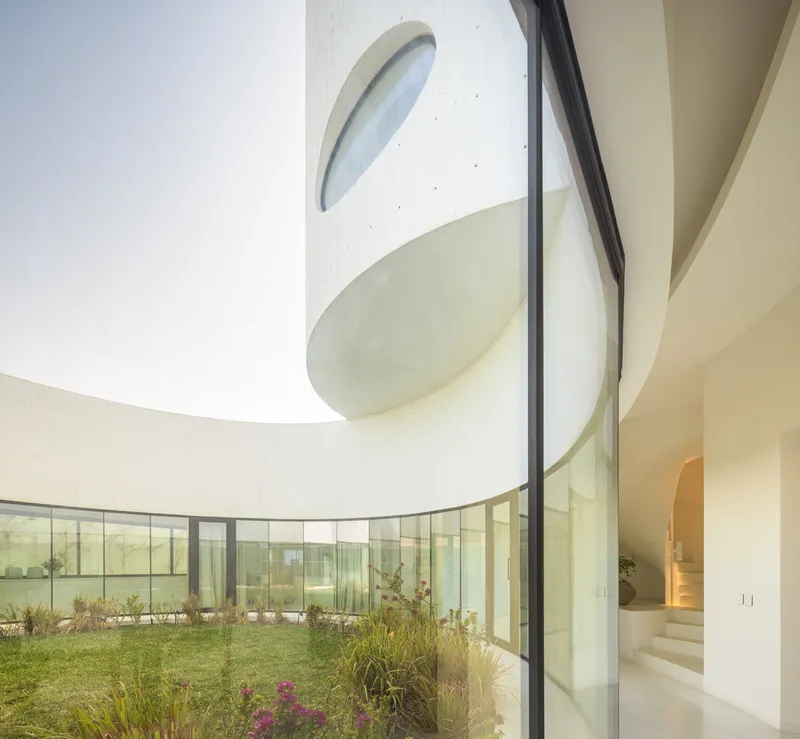 Curving white concrete facade with oval skylights and floor-to-ceiling glazing framing a planted courtyard