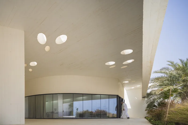 Curved concrete overhang with circular skylights above a person in traditional dress near palm trees