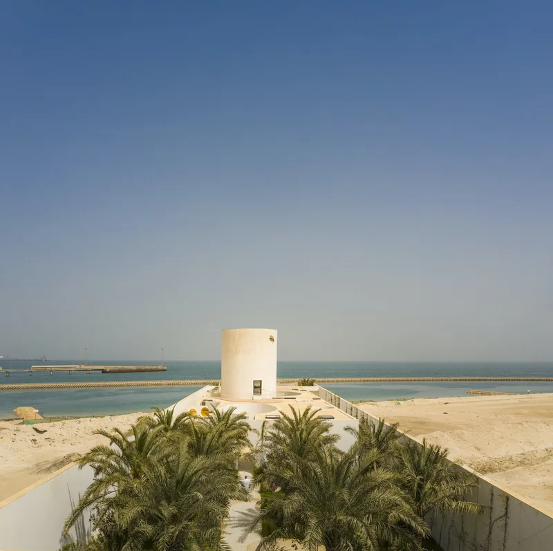 Cylindrical white tower on a coastal promontory surrounded by palm trees and calm turquoise water