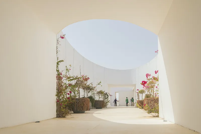 Arched passageway with planted beds of bougainvillea leading to an open courtyard where two figures stand in sunlight