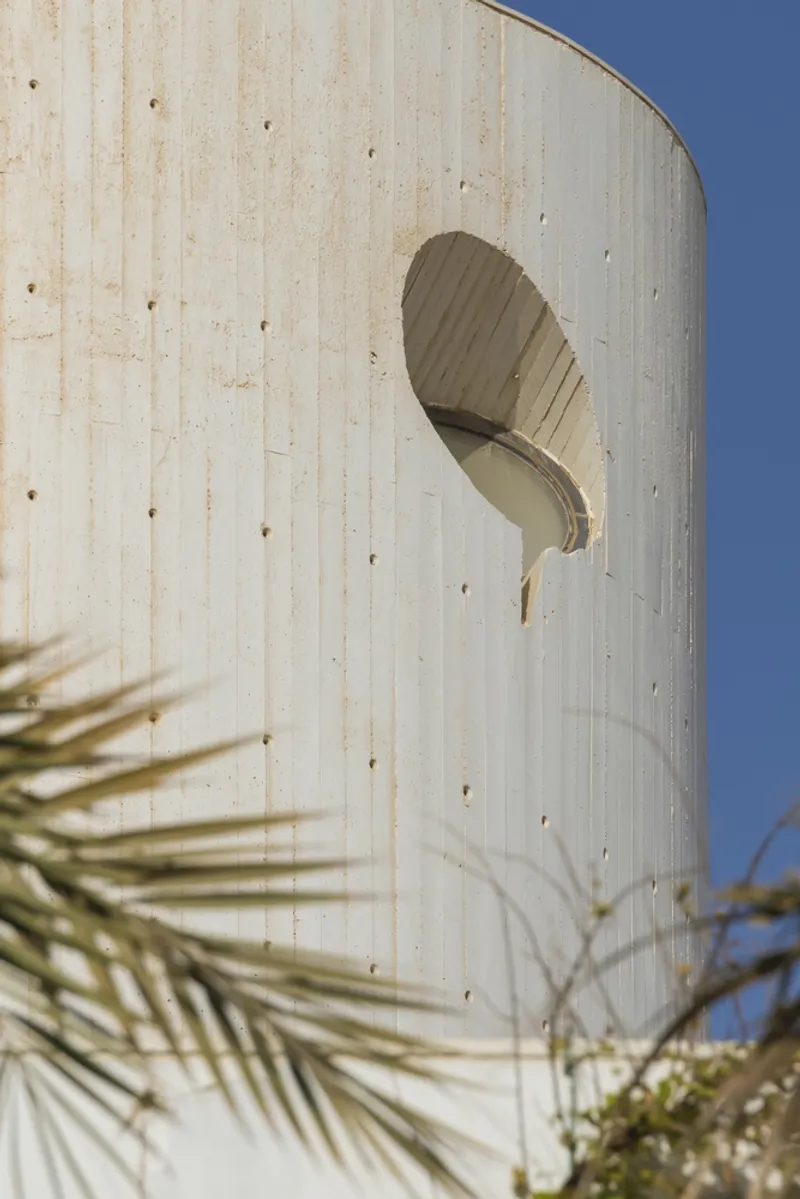 Cylindrical concrete tower with curved window opening and boardmarked formwork pattern against a clear blue sky
