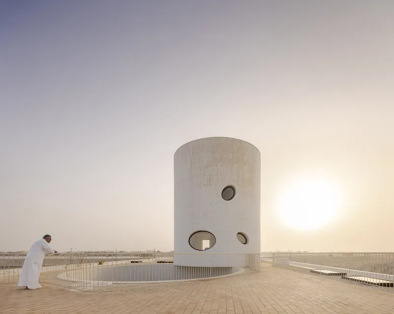 Cylindrical concrete tower with circular openings on a terrace at dusk as a robed figure walks past