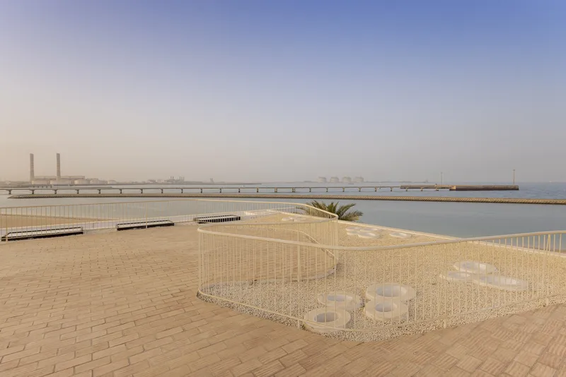 Wire mesh enclosure beside a waterfront promenade with a distant bridge under hazy skies