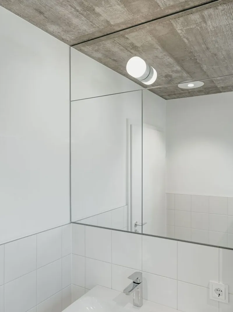 Bathroom mirror reflecting white tiled wall and exposed concrete ceiling with recessed fixtures