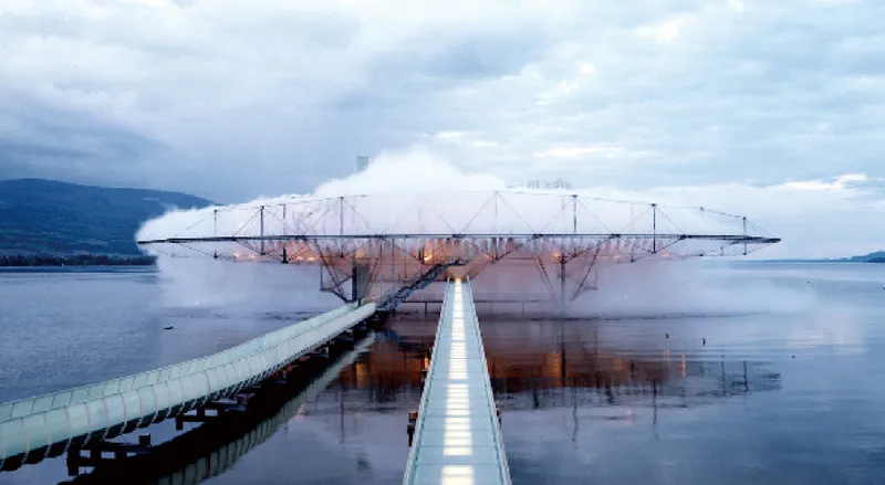 Floating platform with tensile canopy anchored by a pier over calm water under cloudy skies and distant mountains