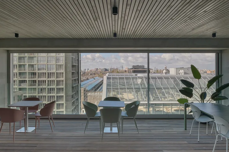 Interior lounge with slatted timber ceiling and floor-to-ceiling glazing overlooking neighboring rooftops under cloudy skies