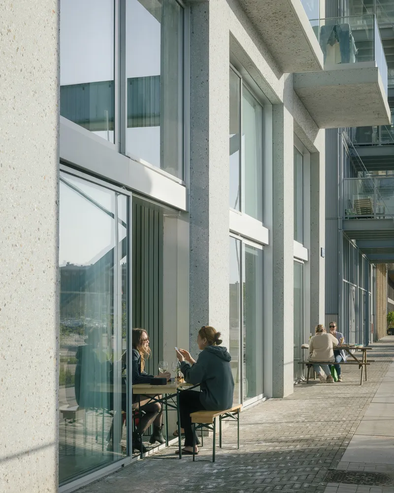 Exterior terrace with tall glazing and concrete columns where residents sit at outdoor tables in afternoon sunlight