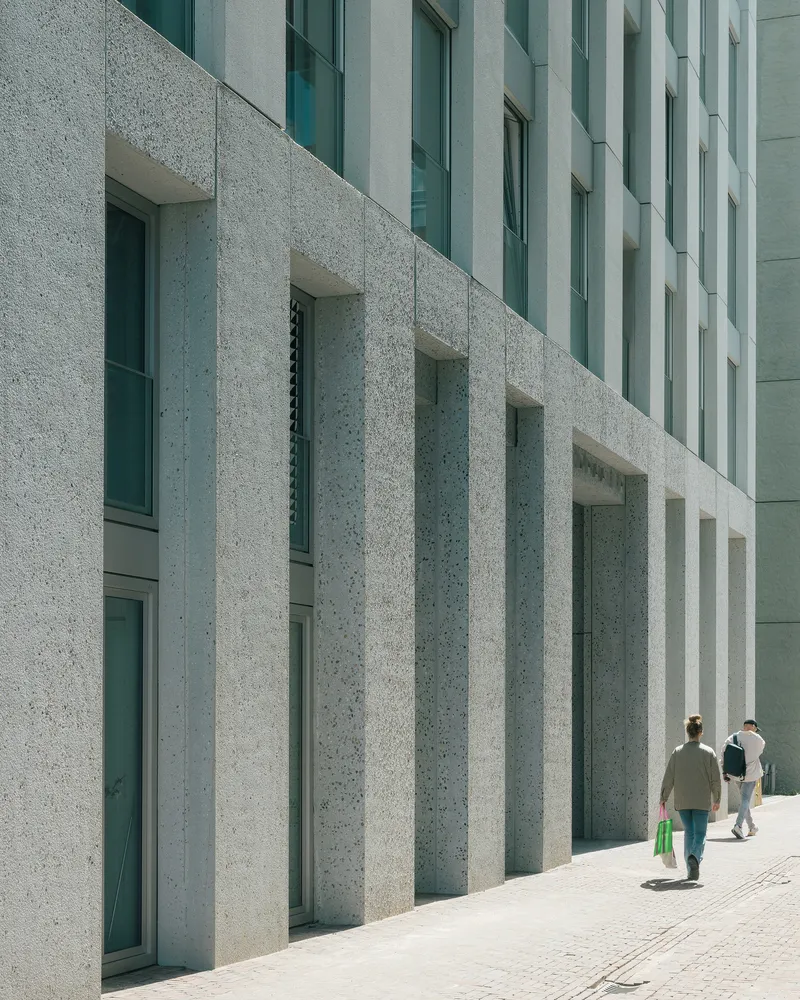 Textured concrete facade with deep vertical fins and recessed windows as pedestrians walk along the pavement