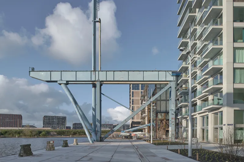 Cantilevered footbridge spanning the waterfront promenade with residential towers and scattered clouds overhead