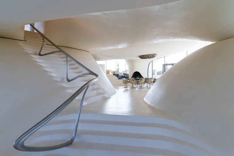 Sculptural white staircase with flowing steel handrail descending into an open living area with fireplace