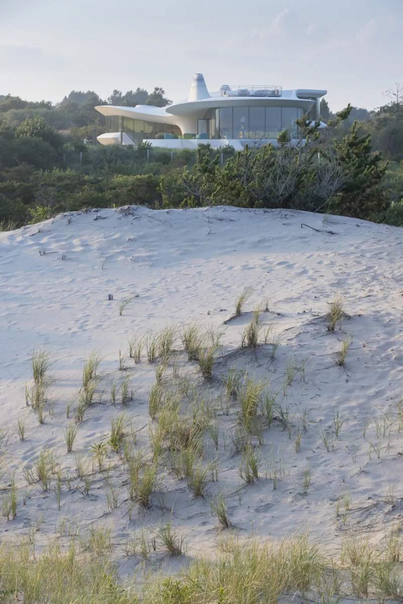 White curved volume with glass walls and cantilevered terraces rising above a sand dune with scattered beach grasses