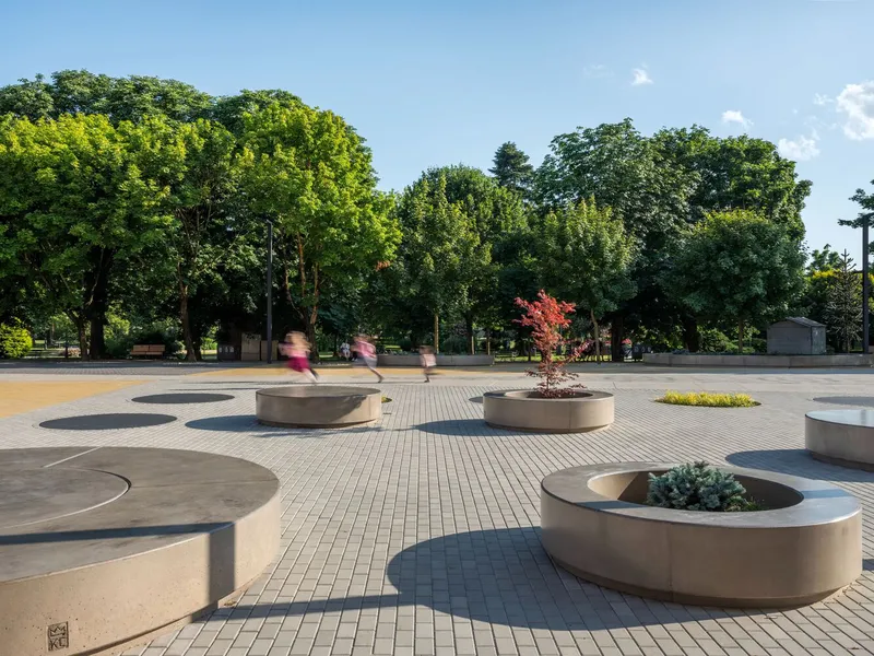 Public plaza with curved concrete planters and paving stones, visitors moving through in afternoon light