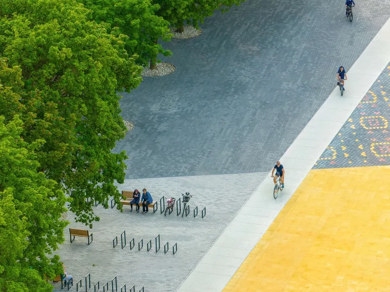 Overhead view of bike path with cyclists passing benches, mosaic paving, and tree canopy