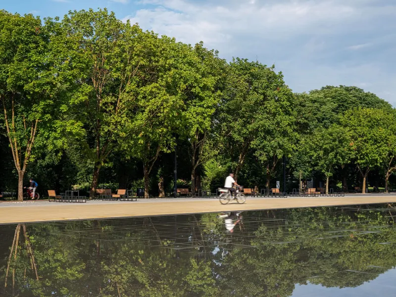 Reflecting pool mirroring a cyclist on the plaza path and tree line beyond