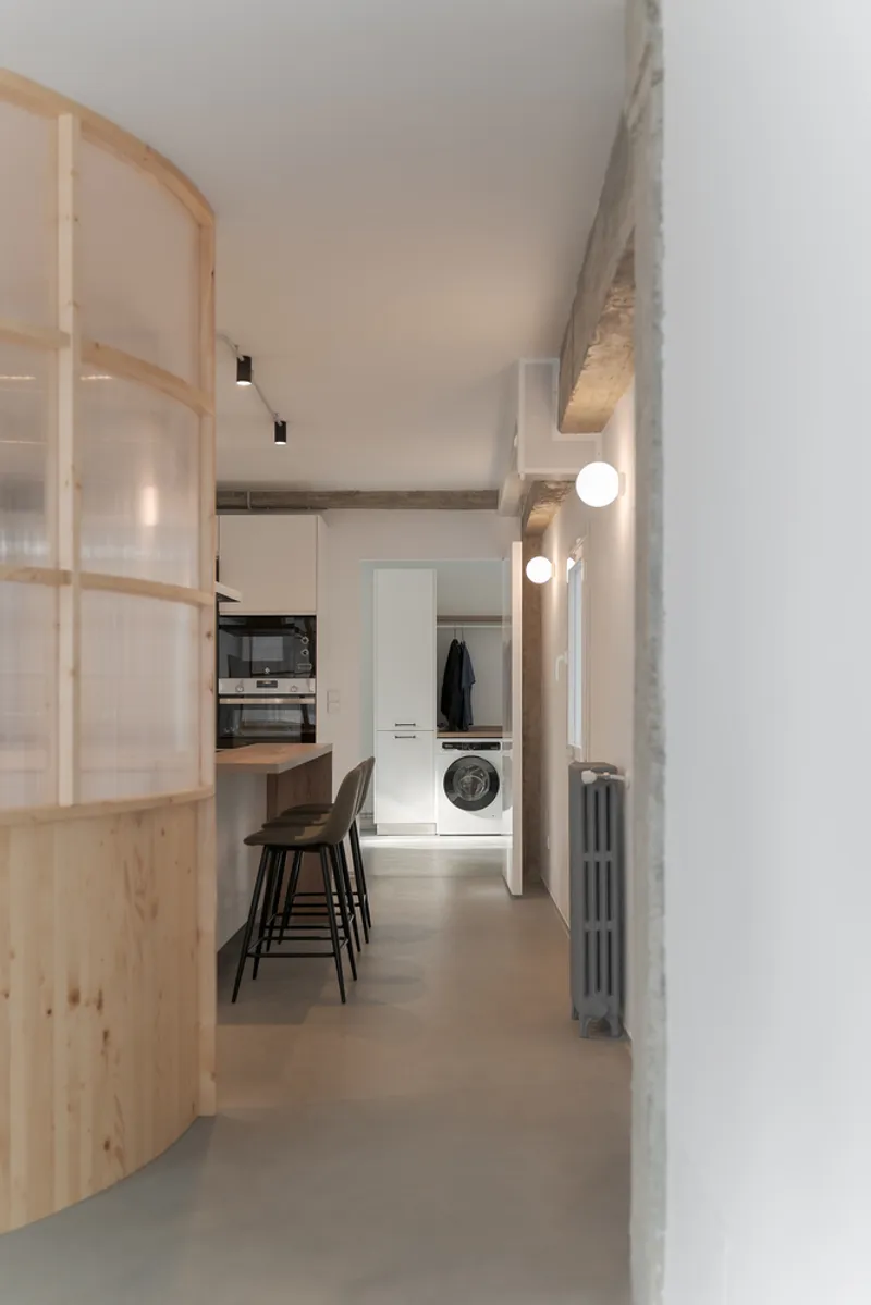 Corridor with curved timber partition and exposed concrete beam leading to kitchen and laundry beyond