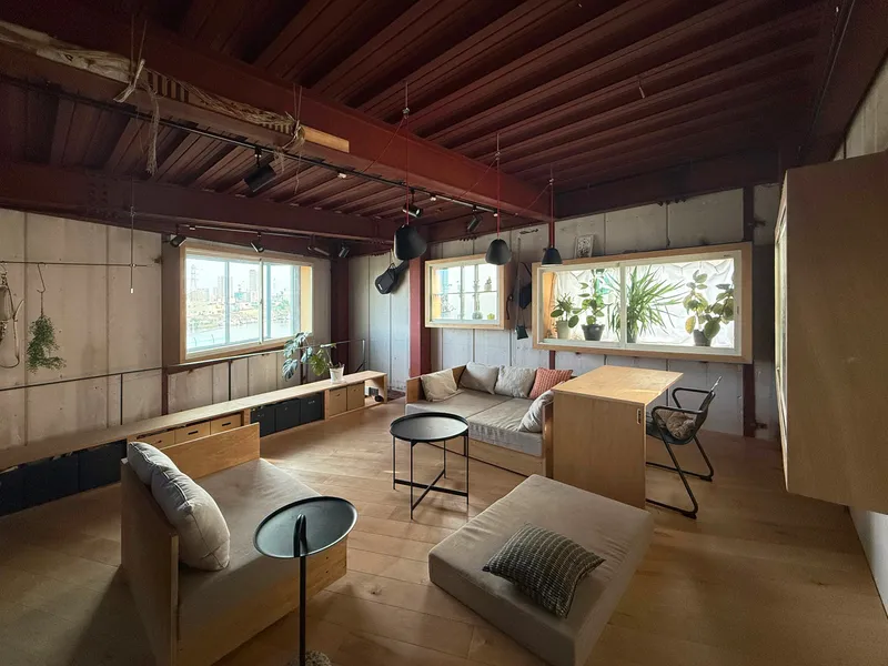 Living space with exposed timber ceiling beams, concrete walls, and windows displaying potted plants