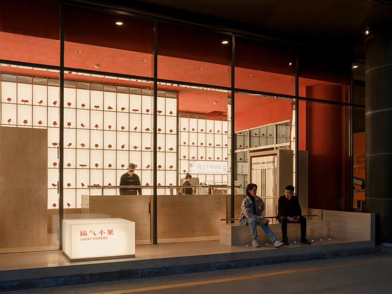 Evening view through storefront glass showing customers at counter and seated on bench at threshold