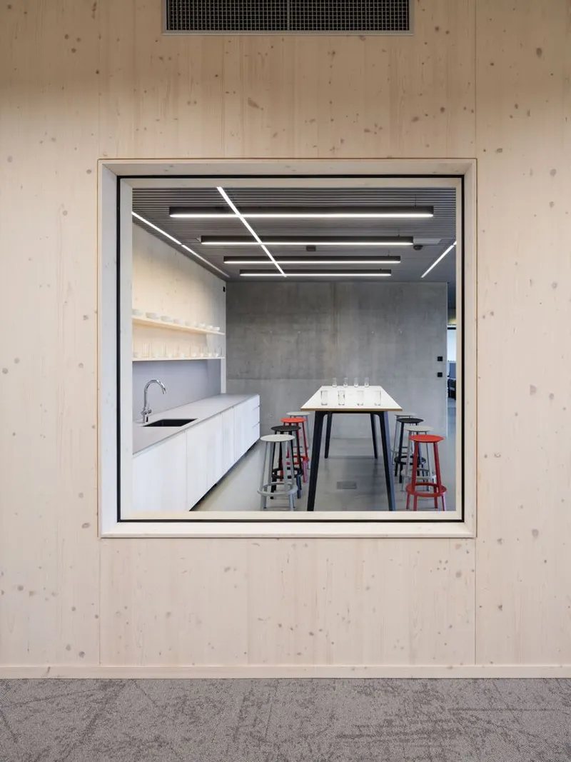 Square window in plywood wall revealing kitchenette with white cabinets and linear ceiling lighting