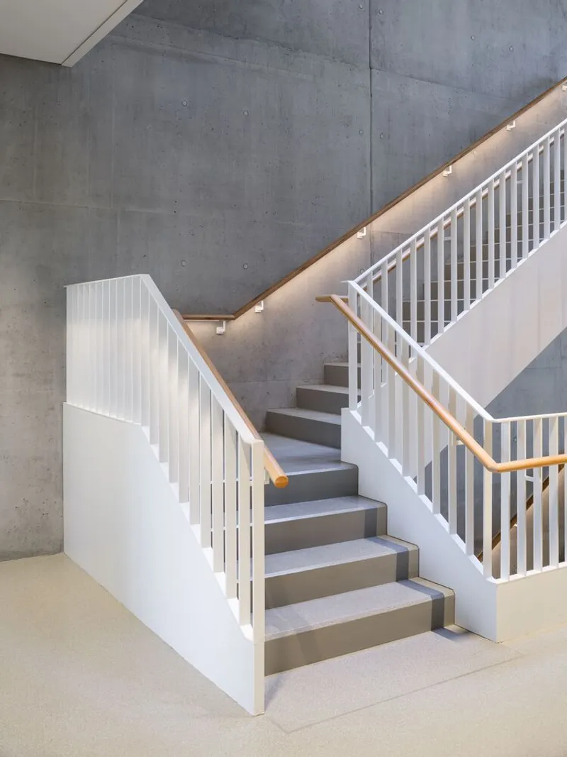White-painted timber staircase with vertical slat railings ascending against a board-formed concrete wall with integrated lighting