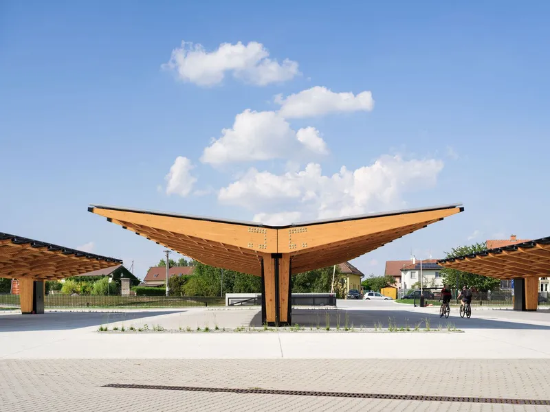 Timber canopy structure with upswept wing forms over a paved plaza with cyclists passing