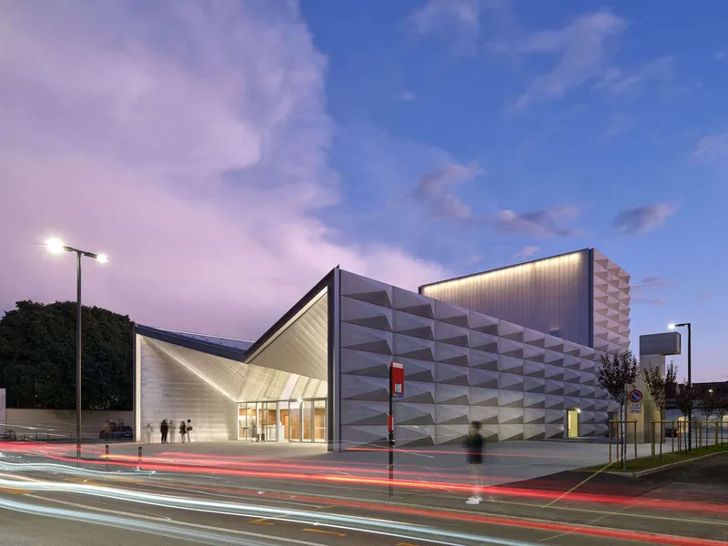 Corner view of the faceted facade and folded canopy entrance with traffic light trails at twilight