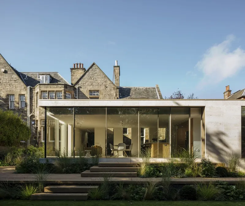 Garden elevation of glazed pavilion extension adjoining stone house with gabled roofs under afternoon sun
