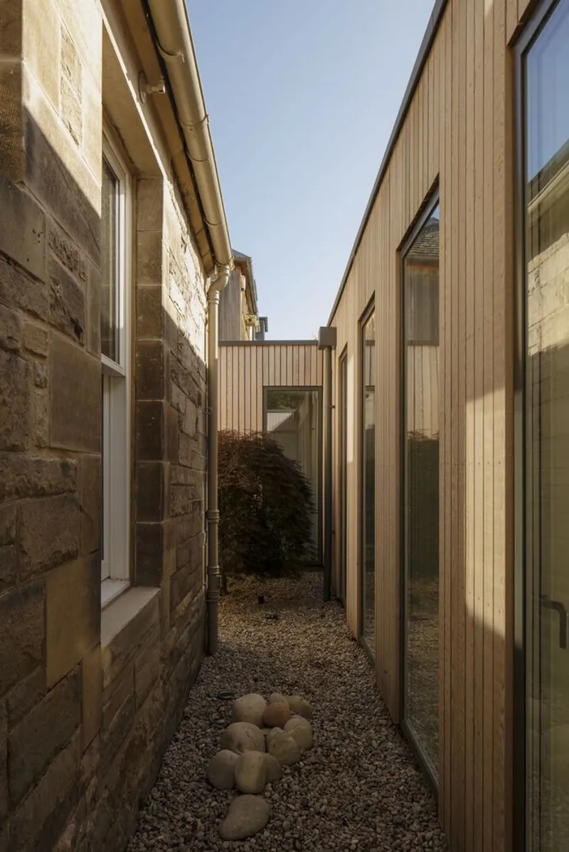 Narrow courtyard between stone masonry wall and vertical timber-clad volume with gravel surface below