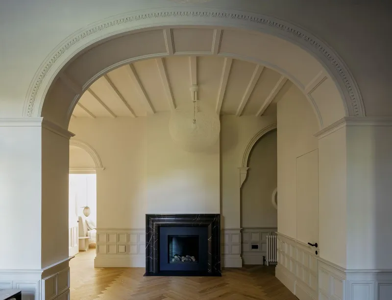 Arched doorway with decorative molding framing an empty room with panel wainscoting and black marble fireplace