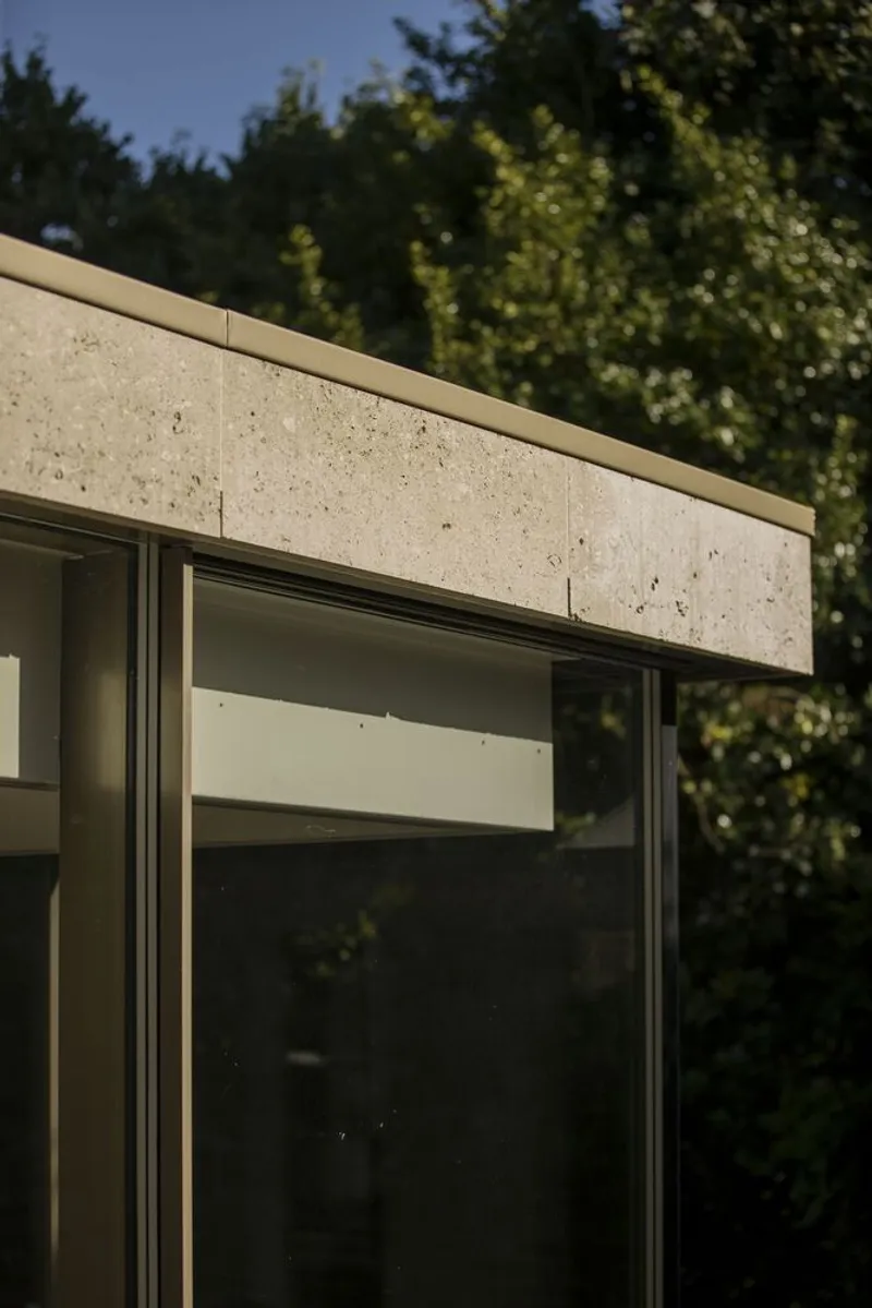 Detail of limestone-clad beam above frameless glass window with forest beyond