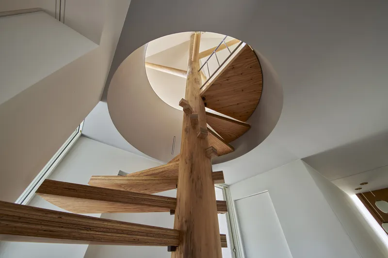 Upward view of timber spiral staircase wrapping around a central tree-like column beneath a circular opening