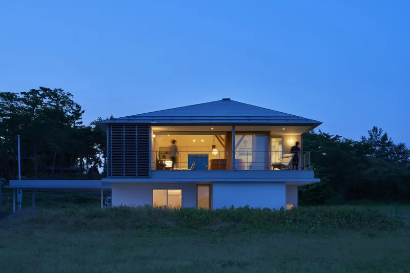 Two-storey house with hip roof and horizontal louvers set in meadow grasses at dusk