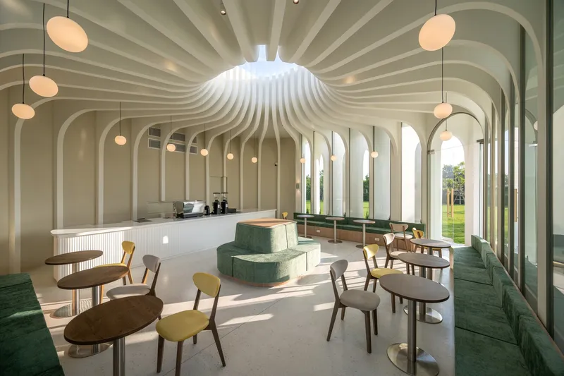 Open cafe space with undulating white ceiling ribs and floor-to-ceiling arched glazing overlooking grass outside